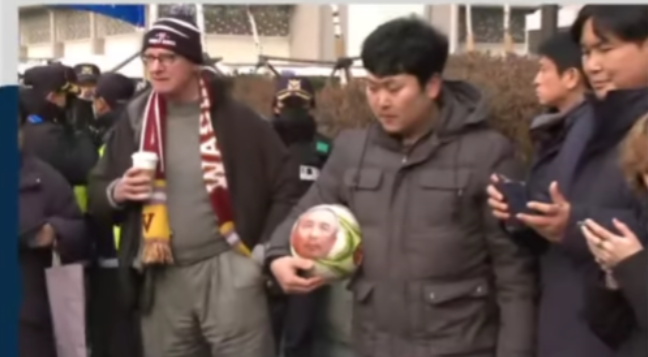 DK, attired in Nationals' World Series beanie, Redskins scarf, hefting coffee cup, covering leftist rally, Seoul, near guy holding soccer ball for decapitated head of Ambassador Harry Harris. Screen grab, Channel A News, Seoul